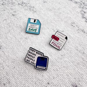 Könnte beinhalten: Drei Emaille-Anstecknadeln mit weißen Rändern. Eine ist eine blaue Diskette mit dem Text "DON'T FORGET TO SAVE". Eine andere ist ein weißes Papier mit dem Text "TRUST ME IT'S FINAL". Die dritte ist ein blaues Buch mit dem Text "THEY WRITE BOOKS ABOUT PEOPLE LIKE YOU".