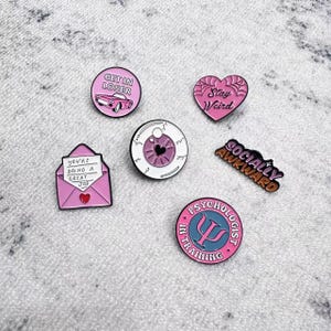 May include: A collection of enamel pins in various shapes and designs. The pins include phrases like "Get in Loser" and "Stay Weird," along with a heart, an eye, an envelope, and a psychologist symbol. The pins are primarily pink, white, and black.