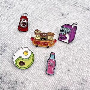 Puede incluir: Colección de pines esmaltados con diseños de comida y bebida. Incluye una botella de ketchup, un perrito caliente con hamburguesa y patatas fritas, una caja de zumo, un aguacate con huevo frito y una botella de zumo rosa. El pin del perrito caliente tiene el texto "LIVE FAST".