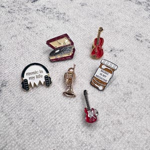 Puede incluir: Colección de pines esmaltados con temática musical. Incluye un tocadiscos, un violín, una guitarra, una trompeta, unos auriculares con el texto "music is my life" y un frasco de pastillas con el texto "music is my medicine".