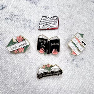 Può includere: Una collezione di spille smaltate con disegni a tema libri. Una spilla presenta un libro aperto con il testo "we lose ourselves in books, we find ourselves there too." Altre spille includono "book nerd", "Sorry I'm booked" e "Cool people read."
