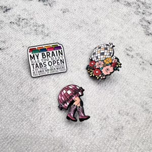 May include: Three decorative pins. One pin features the text "MY BRAIN HAS TOO MANY TABS OPEN" in a rectangular shape. Another pin is a disco ball with flowers. The third pin shows legs wearing cowboy boots.