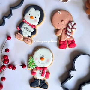 Puede incluir: Galletas navideñas decoradas con forma de pingüino con un cartel de "Christmas", hombre de jengibre y muñeco de nieve con un árbol. Las galletas están decoradas con glaseado y sobre una superficie blanca.