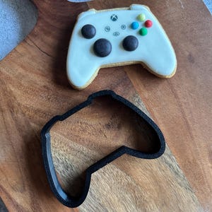 Puede incluir: Una galleta glaseada blanca con forma de mando de videojuego con detalles negros, rojos, verdes y azules. Debajo de la galleta hay un cortador de galletas de plástico negro con forma de mando. Los artículos están sobre una tabla de cortar de madera.
