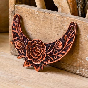 May include: A crescent-shaped copper-toned metal pendant featuring an embossed floral design with a large central rose and smaller roses on each side. The piece has a dark oxidized finish in the recessed areas and two circular holes at the top ends.