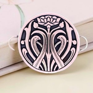 May include: A silver-colored, circular pendant with an Art Nouveau design. The pendant features a black, stylized floral pattern within a silver frame. Two small loops extend from the sides. The pendant is resting on a book with a floral design.