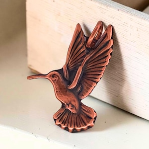 May include: A copper-toned metal pendant shaped like a hummingbird in flight with detailed wing feathers and a long beak. The pendant features a small circular loop at the top for attachment, showcasing a dark, oxidized finish in the recessed areas.