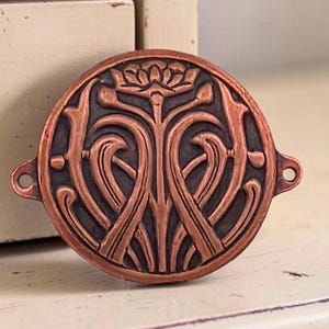 May include: A circular copper-toned metal connector featuring a raised relief design of a stylized flower with symmetrical, curving stems. The piece has a dark recessed background and includes two small loops on opposite sides for attachment.