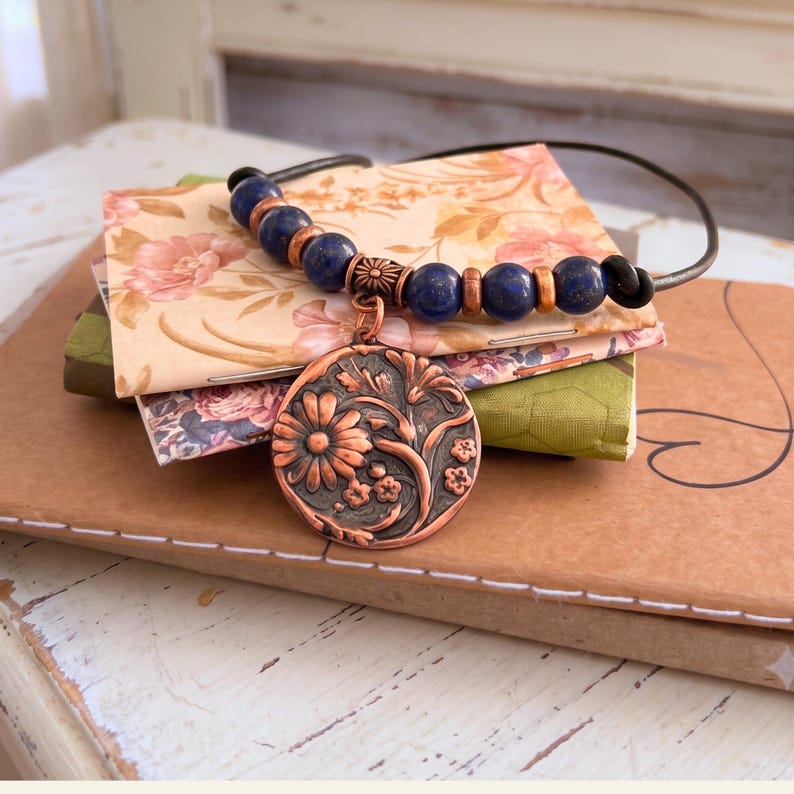 A necklace featuring a black cord strung with multi-colored stone beads, copper-toned spacers, and a central circular copper pendant embossed with a detailed floral design. The pendant displays a raised pattern of flowers and leaves.