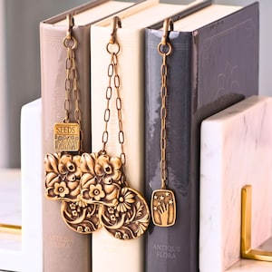 May include: Three metal bookmarks with antique bronze finishes hanging from book spines. The bookmarks feature dangling chains with charms including a square labeled SEEDS, floral embossed squares, circular floral medallions, and a rectangular charm depicting a hand reaching toward stars.