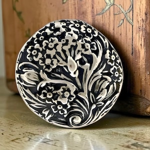 May include: A circular button featuring a high-contrast black and white floral relief pattern. The design displays intricate carved-style details of small blossoms and leaves across the entire surface, creating a textured appearance with deep black recessed areas and raised white floral elements.