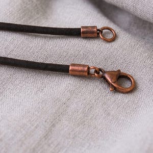 May include: A dark brown cord necklace featuring copper-toned metal findings. One end of the cord is finished with a copper-colored cylindrical crimp and a circular jump ring, while the other end features a matching crimp and a lobster claw clasp.