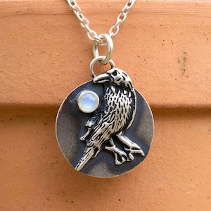 May include: A circular silver pendant featuring a detailed embossed raven standing on a branch, accented with a small round iridescent moonstone. The pendant hangs from a silver chain, showcasing a dark oxidized finish on the metal surface.