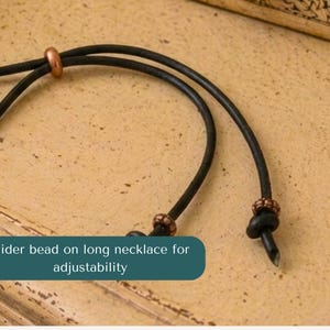May include: A black cord necklace featuring a smooth copper-toned slider bead for length adjustment and two smaller copper-toned beads secured near the knotted ends of the cord. The text slider bead on long necklace for adjustability is visible.
