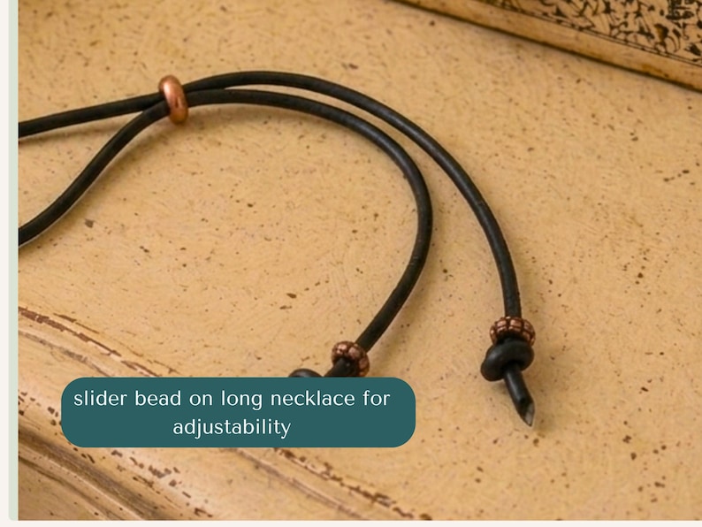 May include: A black cord necklace featuring a smooth copper-toned slider bead for length adjustment and two smaller copper-toned beads secured near the knotted ends of the cord. The text slider bead on long necklace for adjustability is visible.
