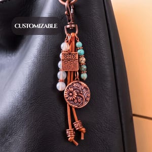 May include: A copper-toned metal bag charm featuring a lobster clasp, a rectangular tag embossed with the word SEEDS, a round floral medallion, and strands of beads in white and turquoise. The charm includes the text CUSTOMIZABLE.