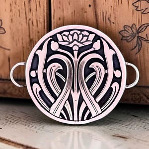 May include: A circular metal pendant featuring a raised, light pink floral and vine relief pattern set against a recessed dark gray background. The piece includes two small, thin metal loops extending from the left and right sides of the disc.
