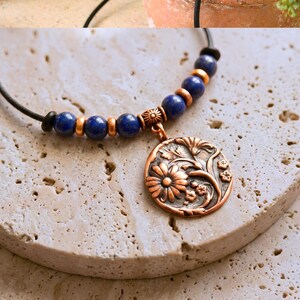 A necklace featuring a black cord strung with multi-colored stone beads, copper-toned spacers, and a central circular copper pendant embossed with a detailed floral design. The pendant displays a raised pattern of flowers and leaves.