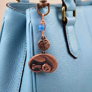 May include: A copper-toned metal bag charm featuring a sleeping cat pendant, a circular disc with an embossed floral pattern, and a single blue bead. The charm is attached to a swivel clasp and hangs from a light blue textured leather handbag.