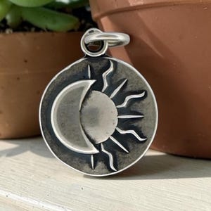 May include: A circular silver pendant featuring a recessed design of a crescent moon and a sun with radiating rays. The metal has a dark, oxidized finish in the grooves, creating a high-contrast look. A silver jump ring is attached to the top.
