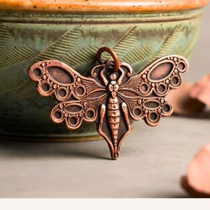 May include: A copper-toned metal pendant shaped like a moth with intricate embossed details on the wings and body. The design features circular patterns and fine lines throughout the surface, with a small circular loop at the top for hanging.