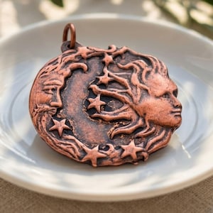 May include: A round copper-toned metal pendant featuring a relief design of a crescent moon face on the left and a sun face on the right, surrounded by several five-pointed stars. The pendant has a small circular loop at the top for hanging.