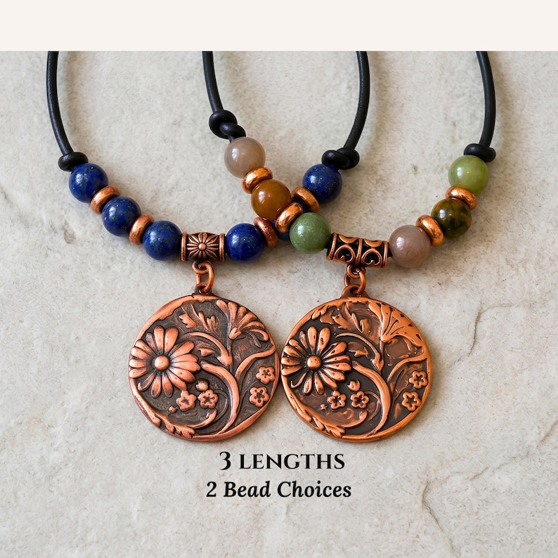 A necklace featuring a black cord strung with multi-colored stone beads, copper-toned spacers, and a central circular copper pendant embossed with a detailed floral design. The pendant displays a raised pattern of flowers and leaves.