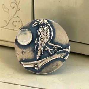 May include: A circular metal knob featuring a raised silver-toned raven perched on a branch against a dark blue background. To the left of the raven is a small, round, iridescent white inlay resembling a full moon. The surface has a polished finish.