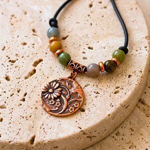 A necklace featuring a black cord strung with multi-colored stone beads, copper-toned spacers, and a central circular copper pendant embossed with a detailed floral design. The pendant displays a raised pattern of flowers and leaves.