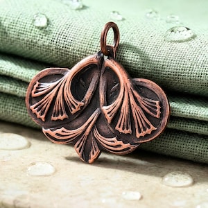 May include: A copper-toned metal pendant featuring a symmetrical design of three stylized ginkgo leaves with dark recessed detailing and a circular jump ring at the top for attachment. The surface has a burnished finish with raised edges.