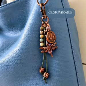 May include: A bag charm featuring a copper-toned dragonfly pendant, a round wheat-embossed charm, and a strand of multi-colored stone beads. The accessory includes a copper lobster clasp and dark green cord accents with the text CUSTOMIZABLE.