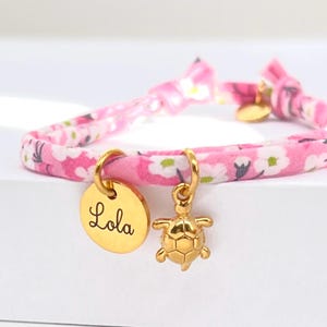 Peut inclure: Un bracelet en tissu rose à motifs floraux avec des breloques dorées. L'une est un disque rond avec le nom "Lola" en cursive, et l'autre est une tortue. Le bracelet est noué avec des nœuds à chaque extrémité.
