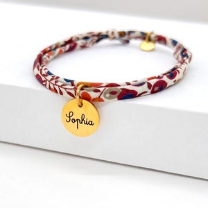 Peut inclure: Un bracelet en tissu avec un motif floral rouge, bleu et crème. Un charm circulaire doré est gravé du nom "Sophia". Le bracelet a un fermoir doré et un petit charm doré.