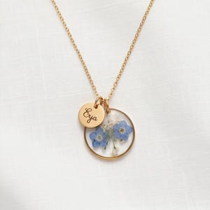 Peut inclure: Un collier doré avec deux pendentifs. L'un est un petit charme rond avec le nom "Eya" gravé. L'autre est un charme circulaire transparent contenant des fleurs bleues et des accents blancs.