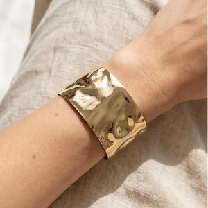 Peut inclure: Un large bracelet manchette doré avec une texture martelée. Le bracelet est porté au poignet, et la lumière se reflète sur la surface texturée. Le bracelet est un bijou de caractère.