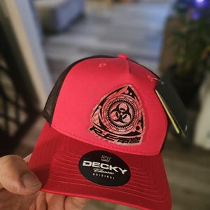 Könnte beinhalten: Rote und schwarze Baseballkappe mit Mesh-Rückseite. Auf der Vorderseite befindet sich ein dreieckiges Emblem mit einem Biohazard-Symbol und dem Wort "REBELS". Die Kappe hat einen gebogenen Schirm und ein Etikett "DECKY Classic Original".