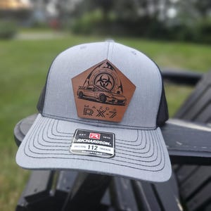 Op de afbeelding: Grijze en zwarte truckerpet met een bruin leren patch met een Mazda RX-7 auto-ontwerp en een biohazard-symbool. De pet heeft een gebogen klep en een verstelbare band. Het merklabel van Richardson is zichtbaar.