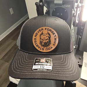 May include: A black trucker hat with a gray mesh back and a curved brim. A circular leather patch features a dog graphic and the text "NEVER FORGET WHY YOU STARTED." The hat has a Richardson 112 tag.