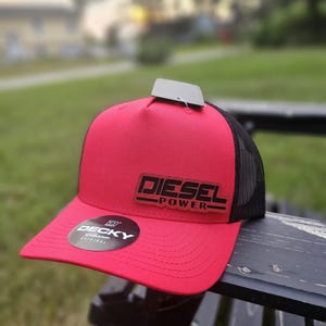May include: Red trucker hat with a black mesh back and a curved brim. The hat features the text "DIESEL POWER" in black lettering on a rectangular patch. A round "DECKY" sticker is on the brim.