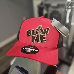 May include: Red and black trucker hat with the words "BLOW ME" in brown and black lettering. The hat has a mesh back and a curved brim. A black tag hangs from the side. The hat has a "Decky" logo.