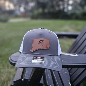 May include: A grey and white trucker hat with a leather patch showing the outline of Connecticut and the letters "CT" with "EST. 1785". The hat is resting on a black wooden chair.