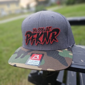 May include: A grey flat bill snapback cap with a camouflage brim. The cap features red and black text that reads "YO SOY DE PFKNR". A white and red Richardson tag is on the brim.