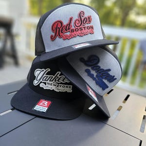 May include: Three baseball caps stacked. The top cap is grey and black with "Red Sox Boston" in red script. The middle cap is grey and black with "Dodgers" in blue. The bottom cap is black with "Yankees New York" in white.