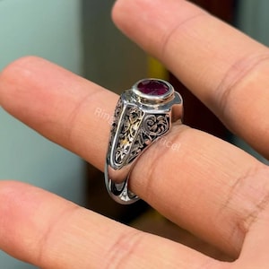Könnte beinhalten: Ein silberner Ring mit einem ovalen, tiefroten Edelstein. Der Ring hat ein verziertes, graviertes Design an den Seiten mit goldenen Akzenten. Der Ring wird an einem Finger gezeigt.