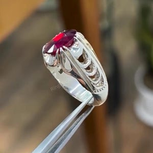 May include: A silver ring with an oval, deep red gemstone. The ring has an ornate, openwork design on the sides. The gemstone is held in place by multiple prongs. The ring is being held by a pair of tweezers.