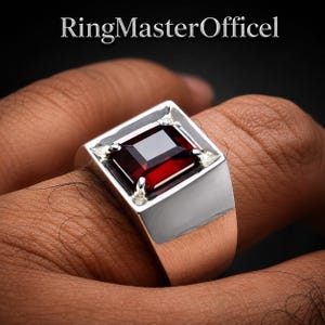 Könnte beinhalten: Ein silberfarbener Ring mit einem rechteckigen, dunkelroten Edelstein in einer quadratischen Fassung. Die Fassung wird durch vier Krappen an den Ecken gehalten. Der Text RingMasterOfficel ist oben vor dunklem Hintergrund sichtbar.