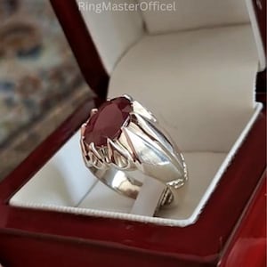 Könnte beinhalten: Ein silberner Ring mit einem großen, oval geschliffenen, tiefroten Edelstein. Der Ring ist in einem dekorativen Silberband gefasst und wird in einer roten Schmuckschatulle präsentiert. Das Design des Rings weist aufwendige Details um den Edelstein auf.