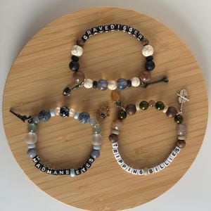 May include: Three beaded bracelets featuring various stone and glass beads. One bracelet displays the text GRAVEDIGGER with a shovel charm. Another shows MADMANS EYES with a hamsa charm. The third features TRIPPING BILLIES with a pineapple charm.