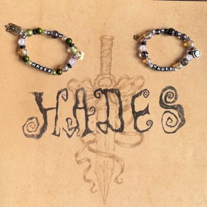 Op de afbeelding: Twee armbanden met kralen met het woord "HADES" gespeld met letterkralen. De armbanden hebben groene, paarse en transparante kralen. Het woord "HADES" staat ook in grote letters eronder, samen met een zwaard.