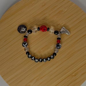 May include: A beaded bracelet spelling "RAW FEAR" with black and white letter beads. The bracelet includes a red skull bead, clear beads, and a silver trainer charm. A black and white circular charm is also included.